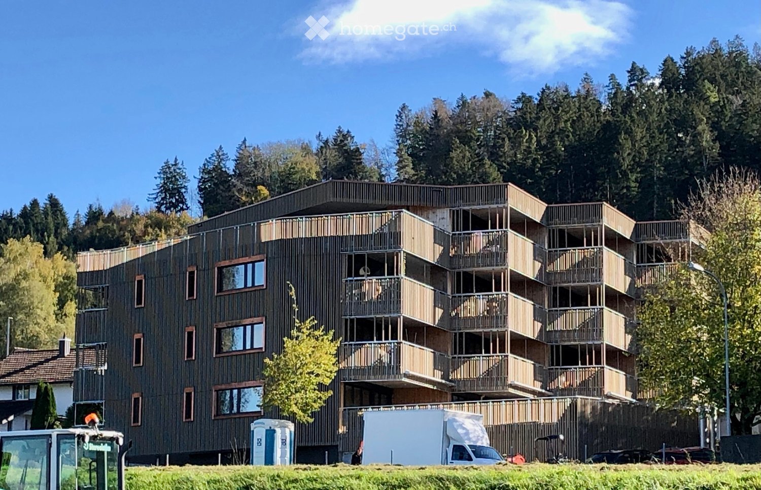 Modern apartment building with balconies, brown exterior, multiple floors, surrounded by trees and grass