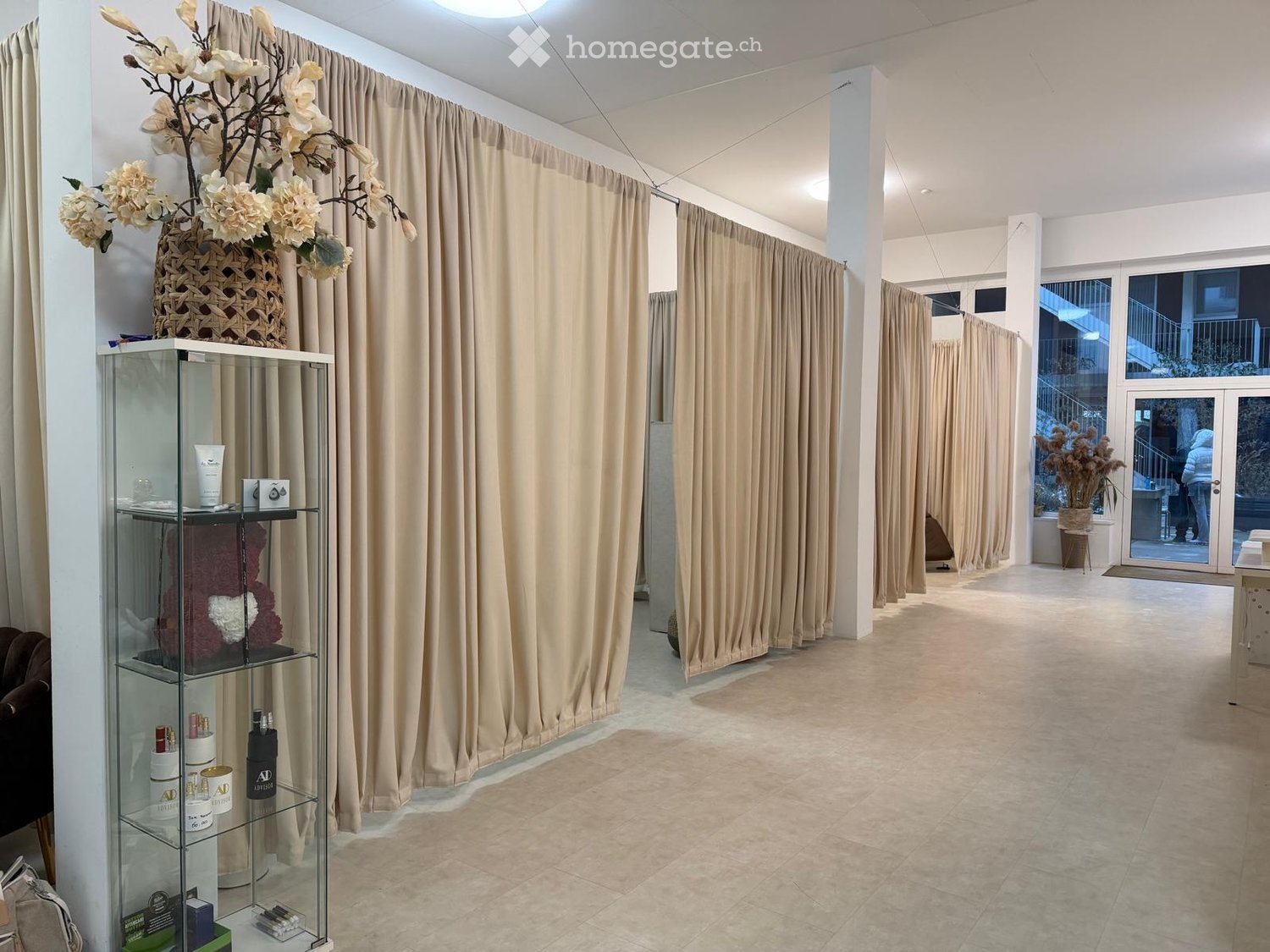 Empty room with beige curtains, tiled floor, glass display case with products, and a flower vase on top.