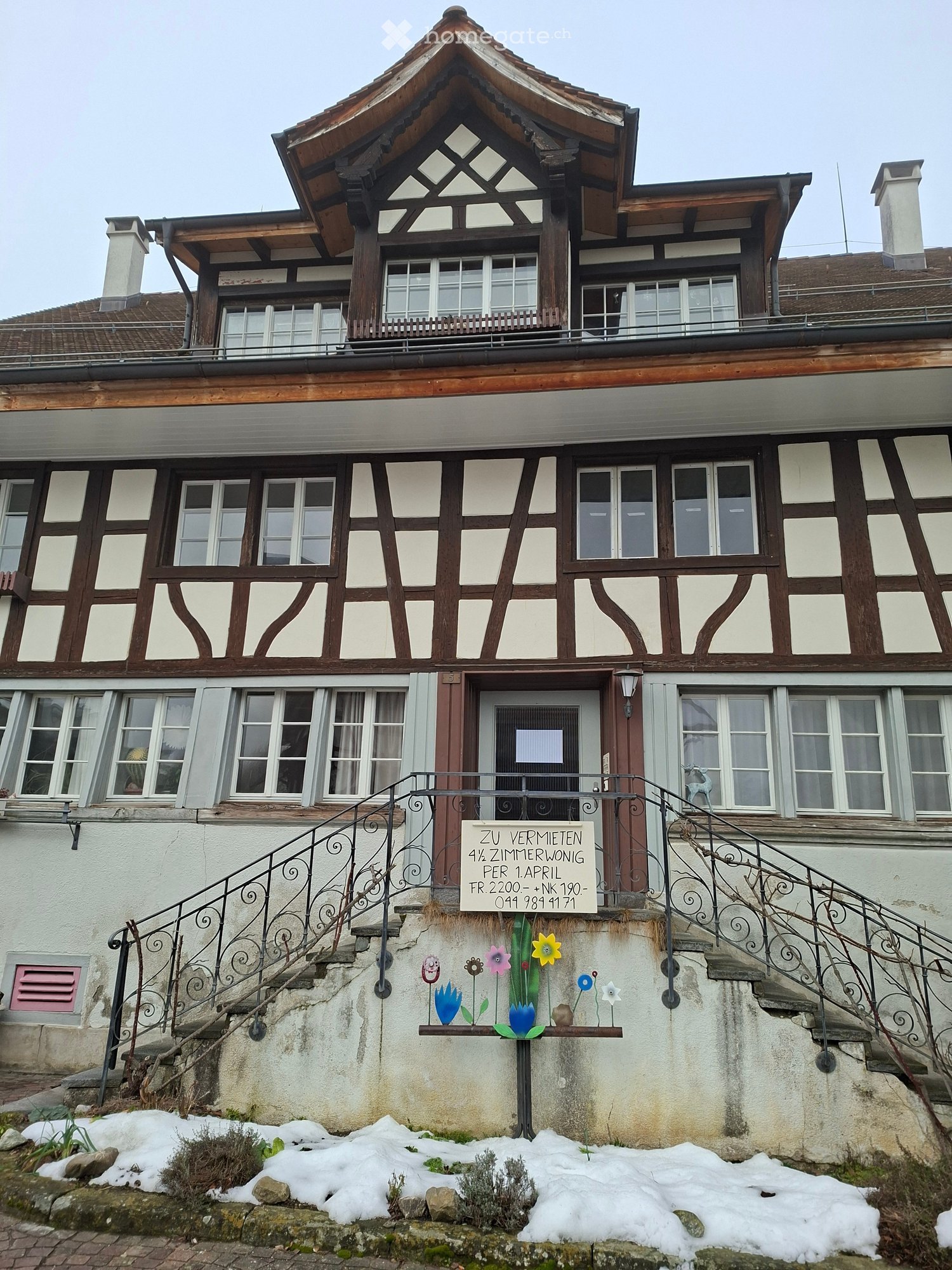 Traditional house with wooden and white walls, multiple windows, a door with a sign, and a staircase leading to the entrance.