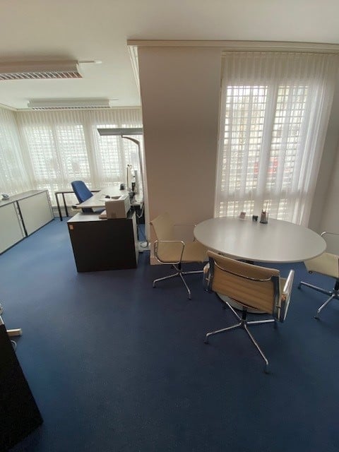 An empty office with a round table, chairs, desks, and windows with white curtains