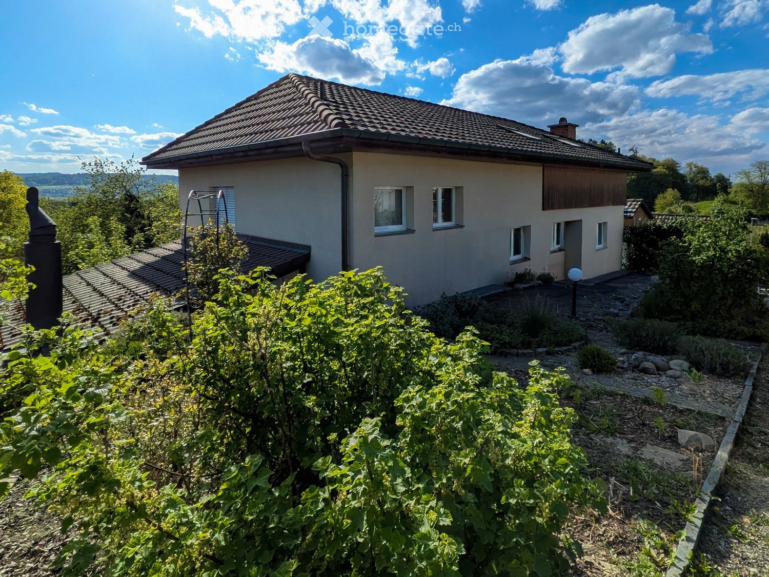 House. Single-story house with a tiled roof, surrounded by a garden with lush greenery and a stone pathway leading to the entrance.