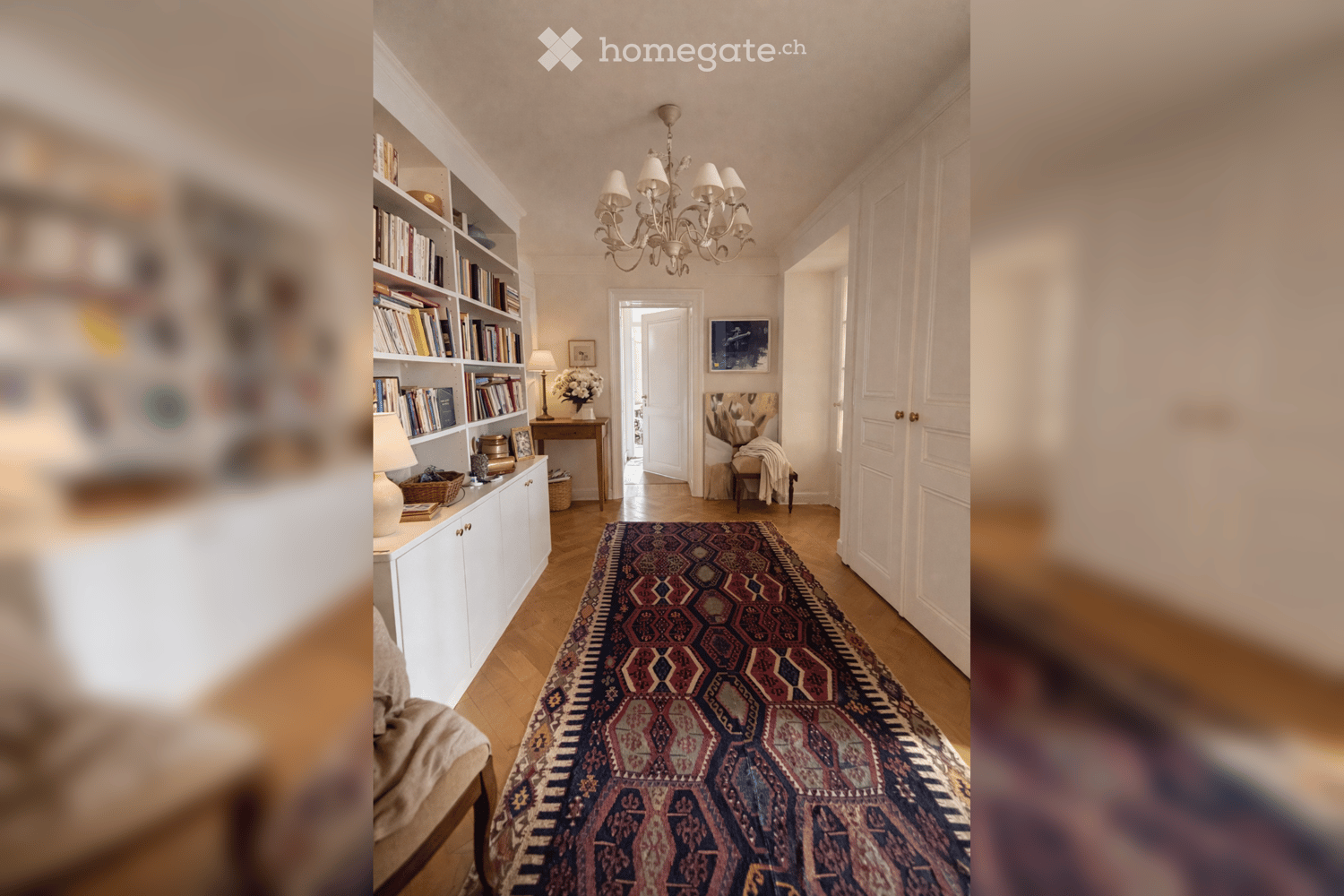 long hallway, wooden floor, chandelier, white cabinets, bookshelves, rug, picture frame, table with flowers, lamp