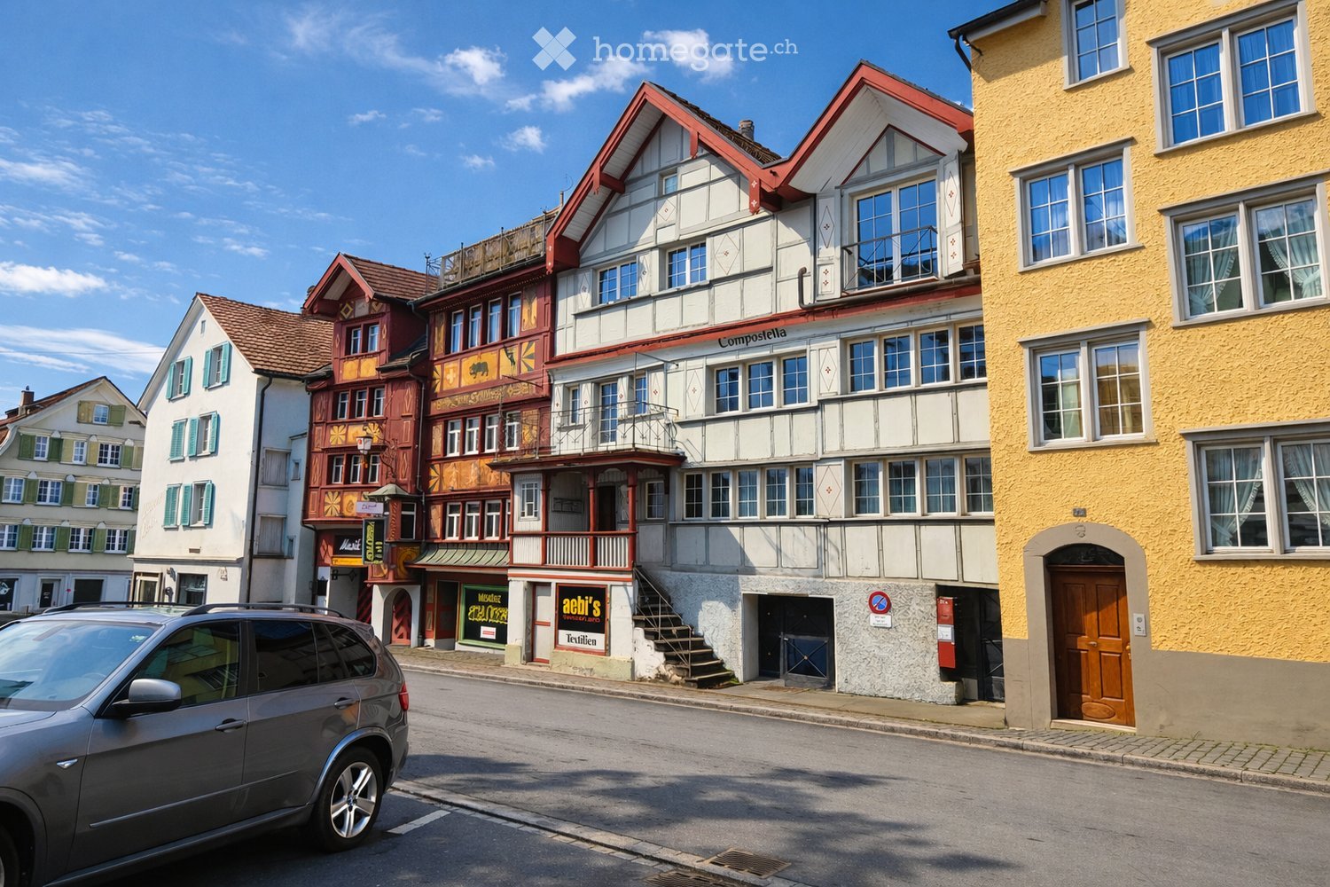 A row of colorful, historic buildings with varied architectural styles, including red and white facades, balconies, and shops at the ground level.
