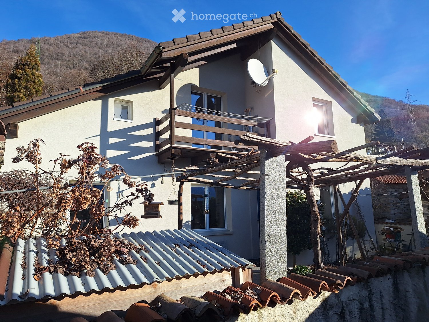 white house, brown roof, balcony, satellite dish, metal roof on the left, plants, trees, mountains in the background
