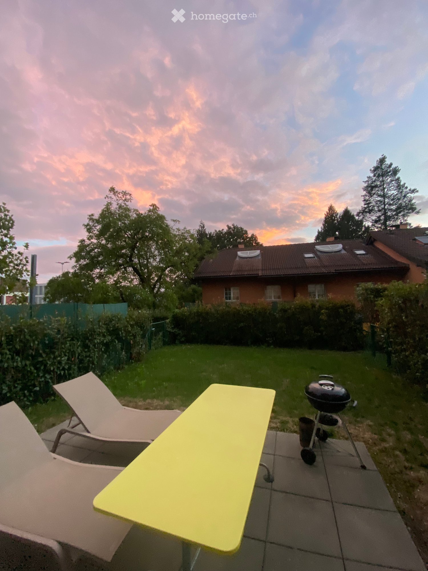 A garden with a yellow table, two lounge chairs, a barbecue, and a house in the background.