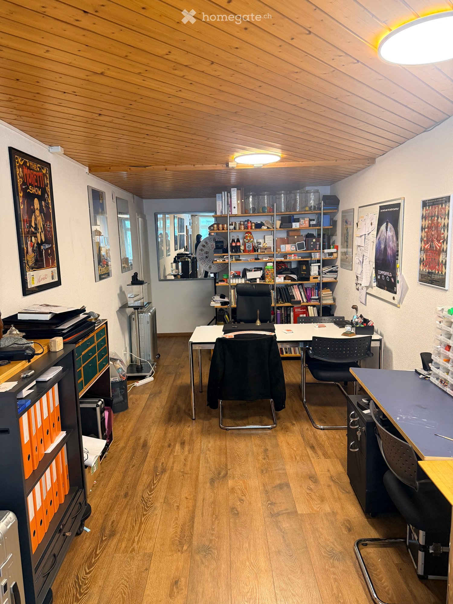 Office room with wooden floor, white walls, ceiling lights, desks, chairs, shelves, and various items