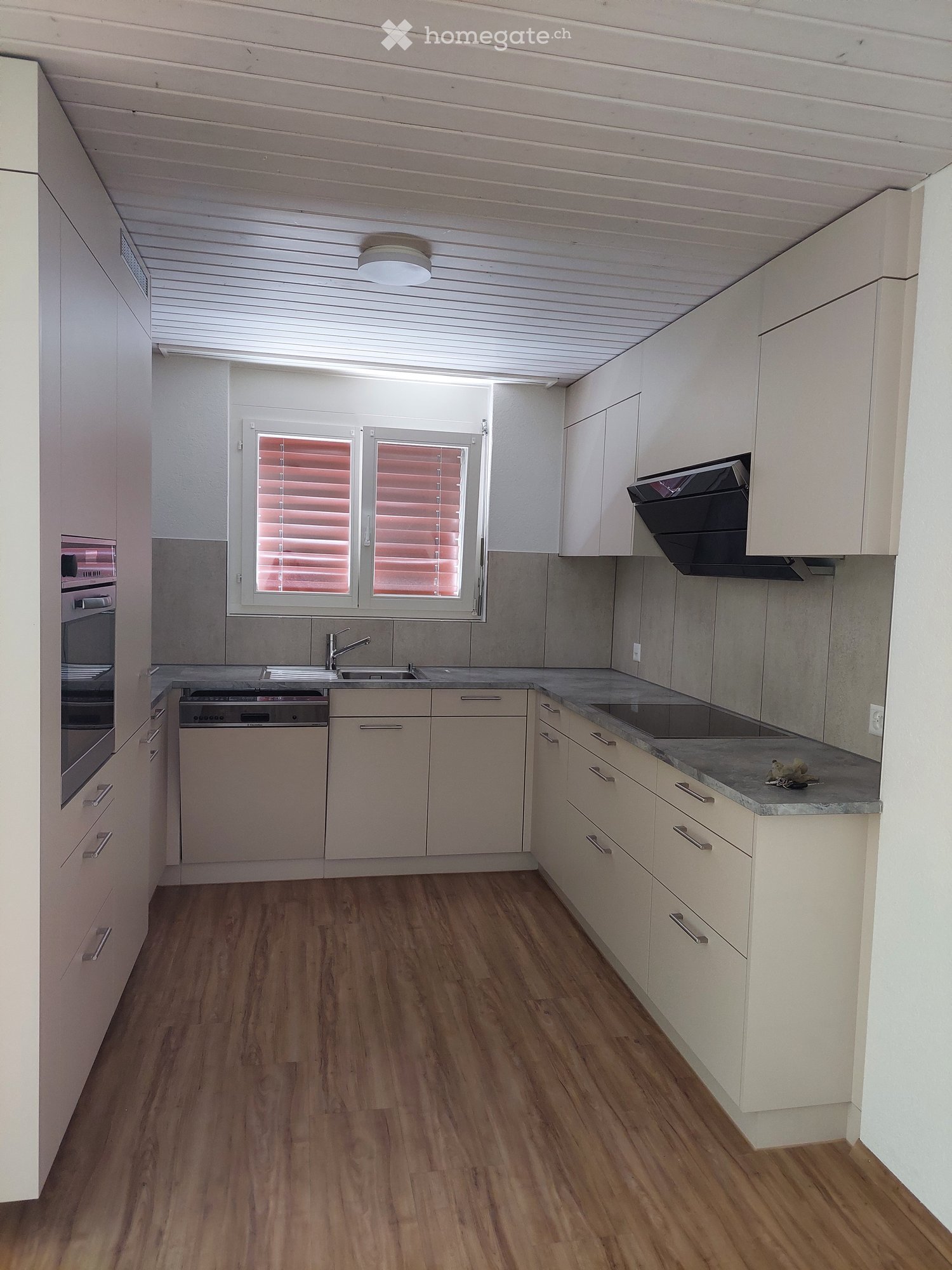 Modern kitchen with light-colored cabinets, wooden flooring, and stainless steel appliances including a sink, oven, and dishwasher. Features a large window with blinds and a range hood above the stove.