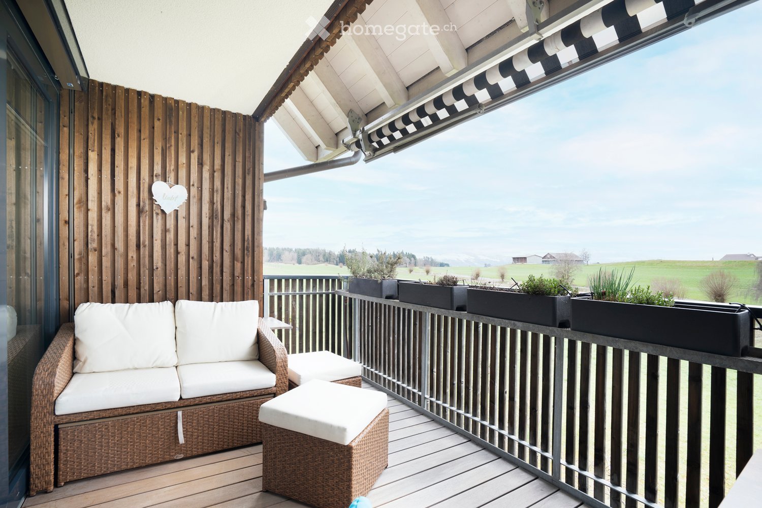 Balcony with wooden wall, couch, ottoman, metal railing, plants, view of the field