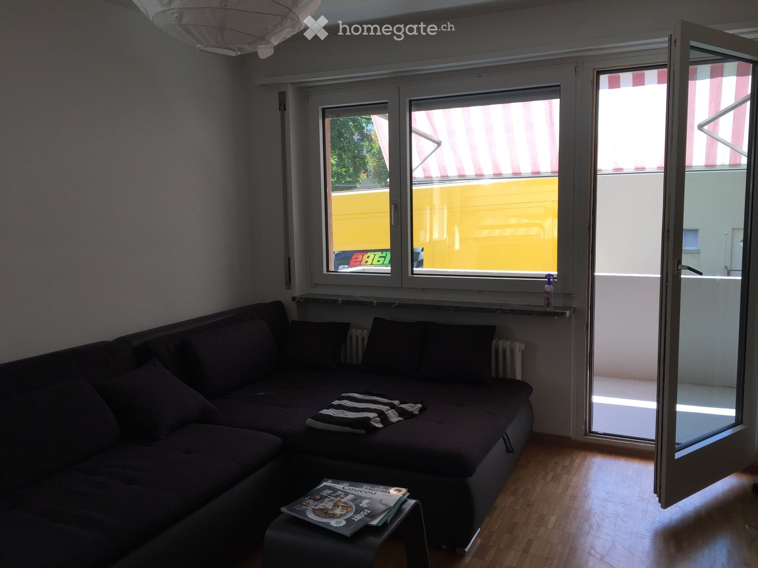 L-shaped black sofa, coffee table with magazines, open door to balcony, white walls, wooden floor