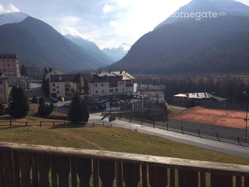 Building with multiple floors, surrounded by mountains, tennis court, parking lot, and trees