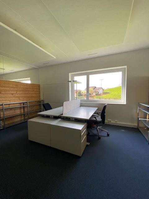 An empty office with a desk, chairs, and a window. The office has a wooden wall, a white ceiling, and a blue carpet. There is a metal shelf on the left side of the room.