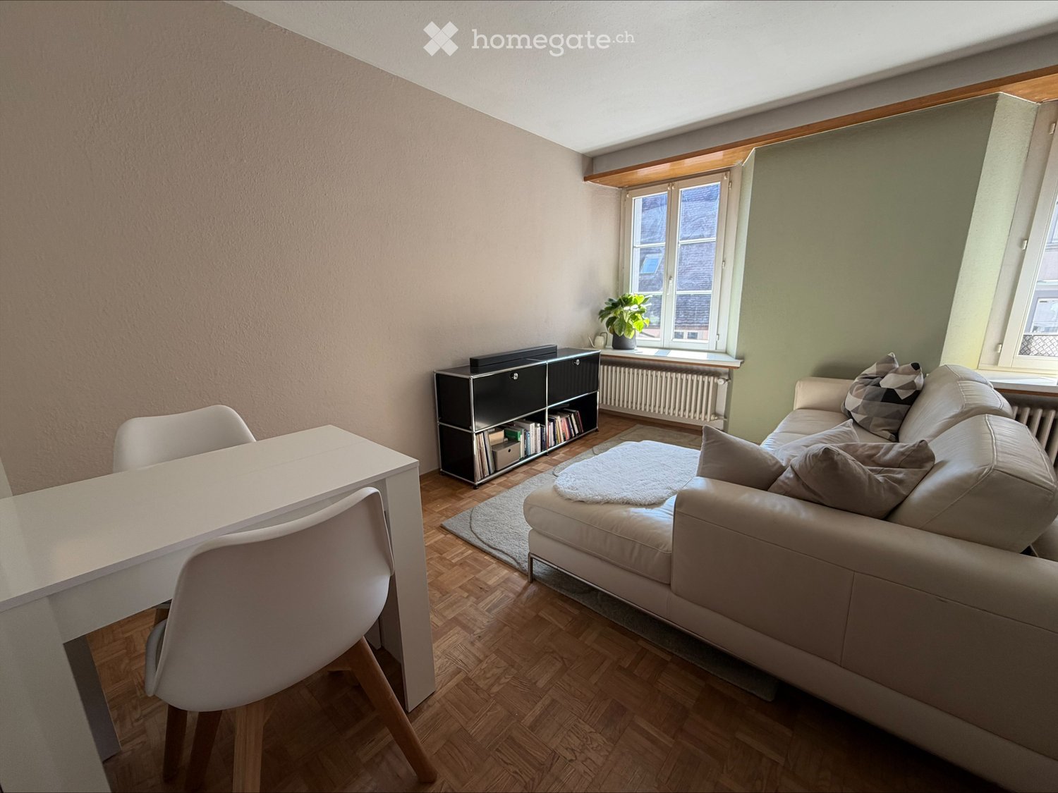 living room with white couch, white desk, white chairs, wooden floor, black cabinet, books, plant, radiator, windows