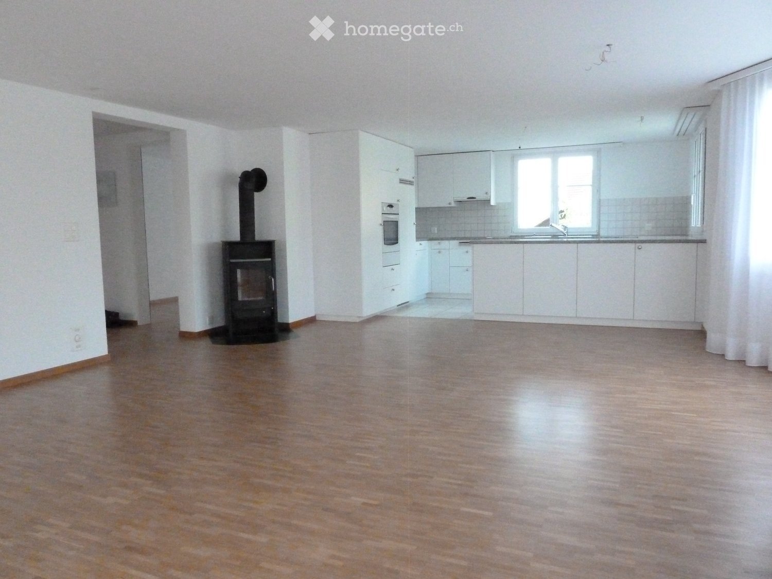 Empty room with wooden flooring, white walls, and a fireplace. The room includes a kitchen area with white cabinets and a sink.