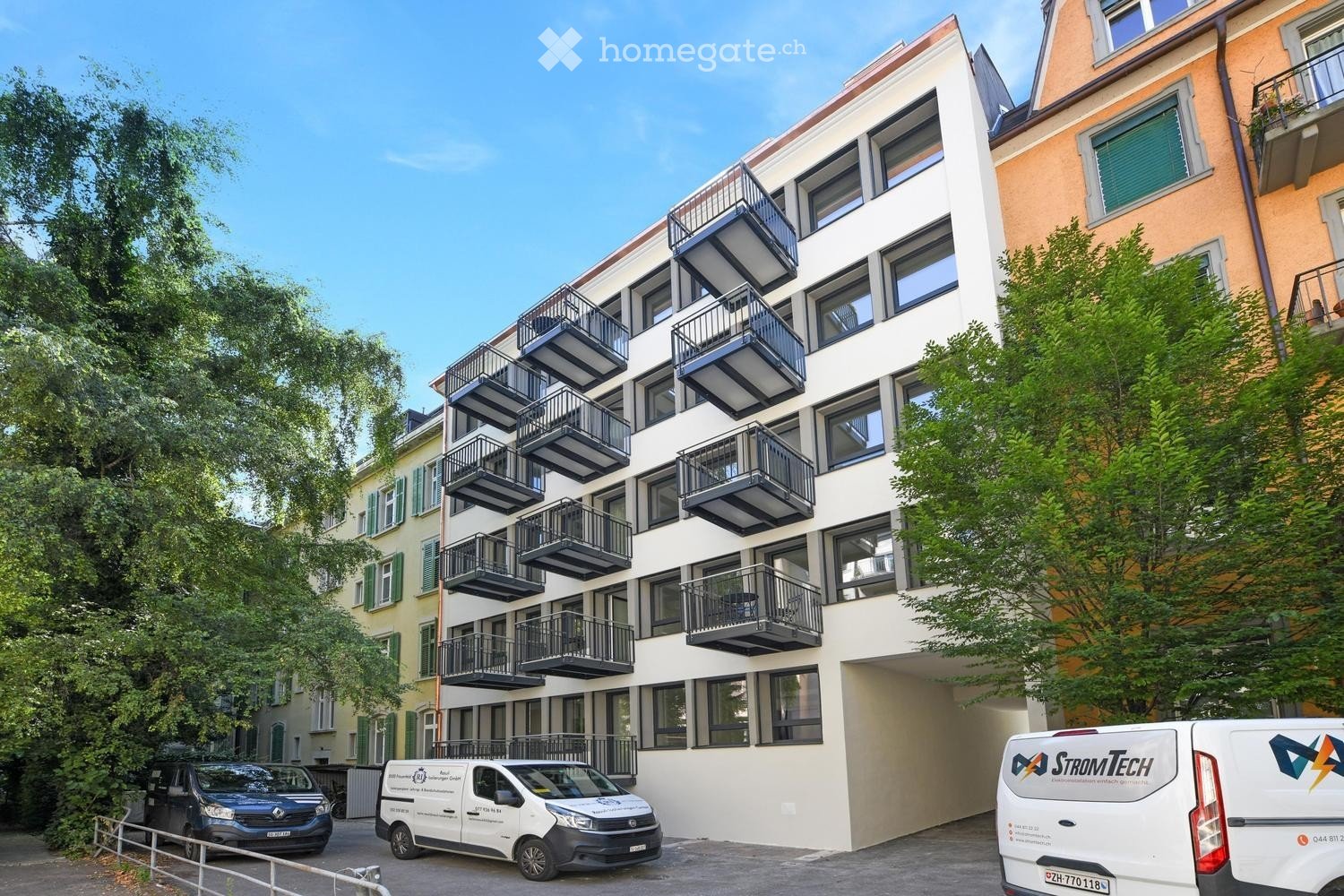 Modern apartment building with balconies, white exterior, multiple floors, trees in front