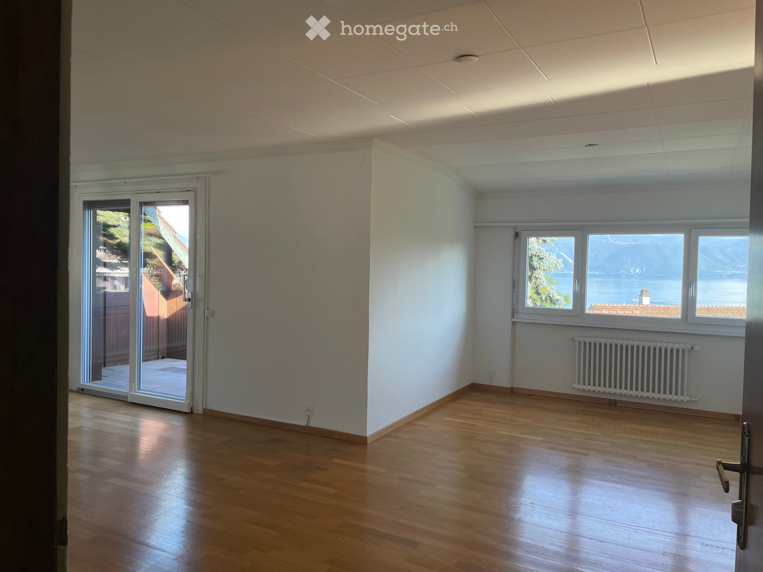Empty room with wooden flooring, white walls, large windows, and a sliding glass door leading to a balcony.