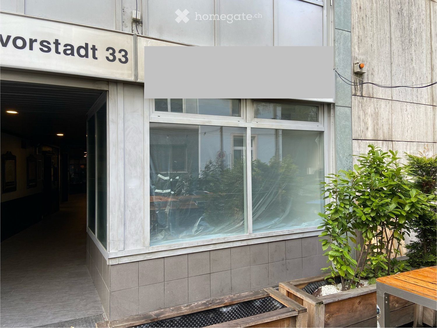 Exterior of a building with a sign reading 'Vorstadt 33', glass windows, wooden planters, and a wooden table.