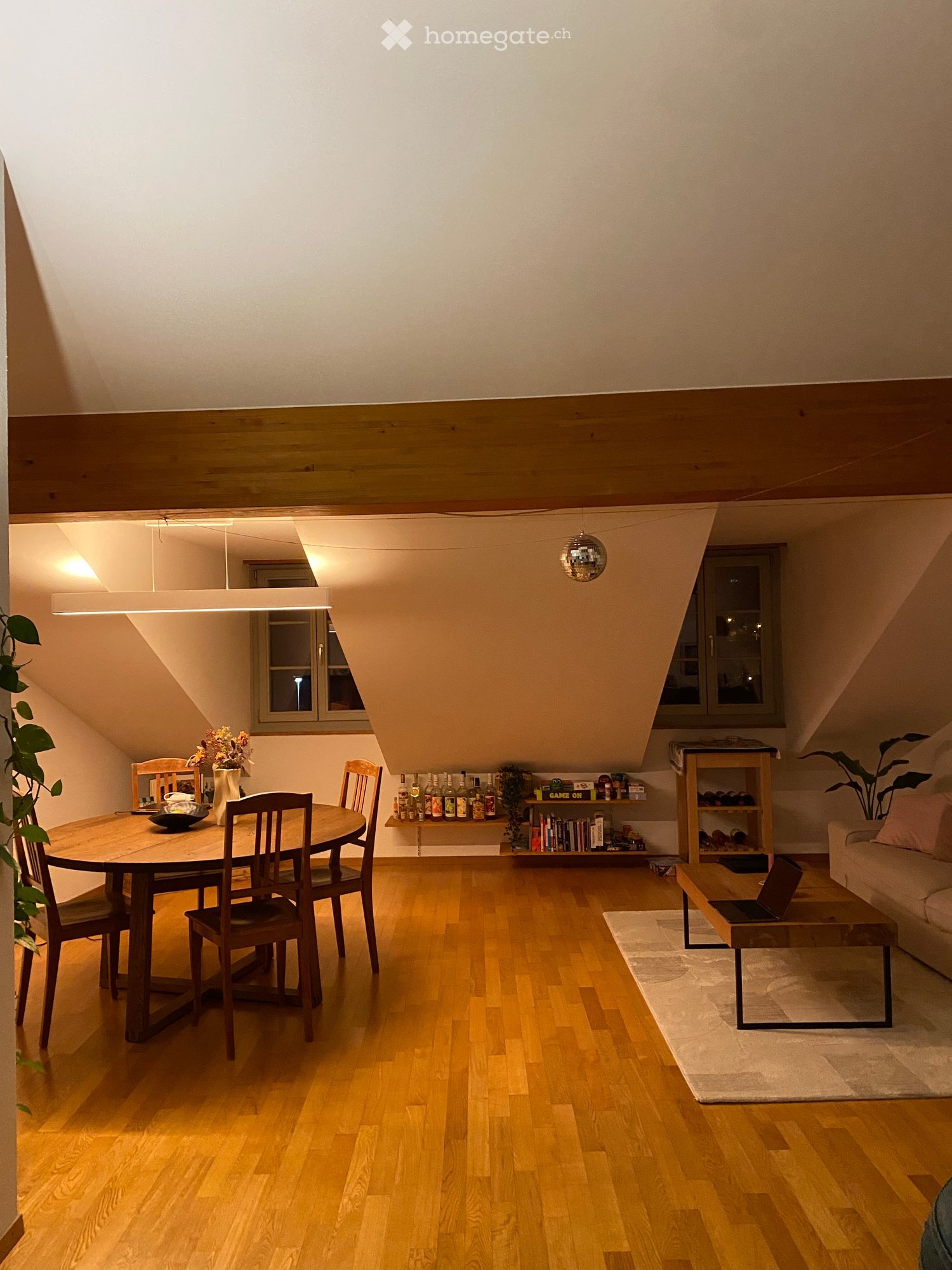A cozy attic room with wooden floors, a round dining table, wooden chairs, a wooden coffee table, a couch, a shelf with books and bottles, and a plant.