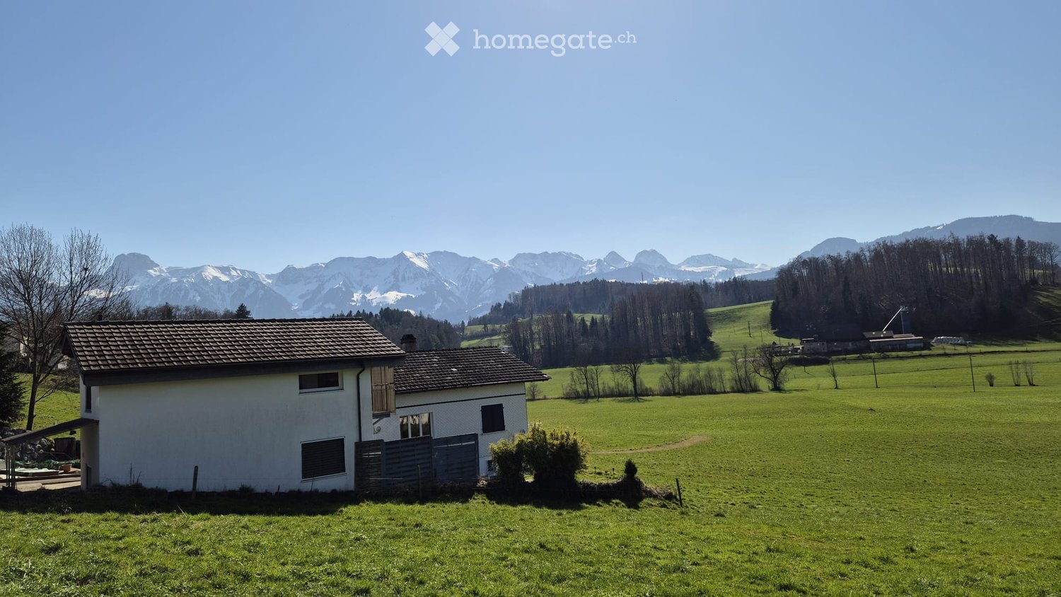 Single story house, brown roof, white walls, surrounded by green field, mountains in the background
