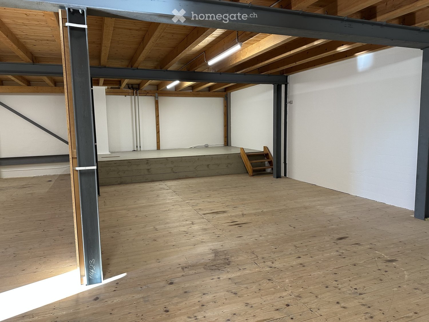 Empty room with wooden floor, white walls, wooden ceiling, metal beams, stage, stairs, and lights