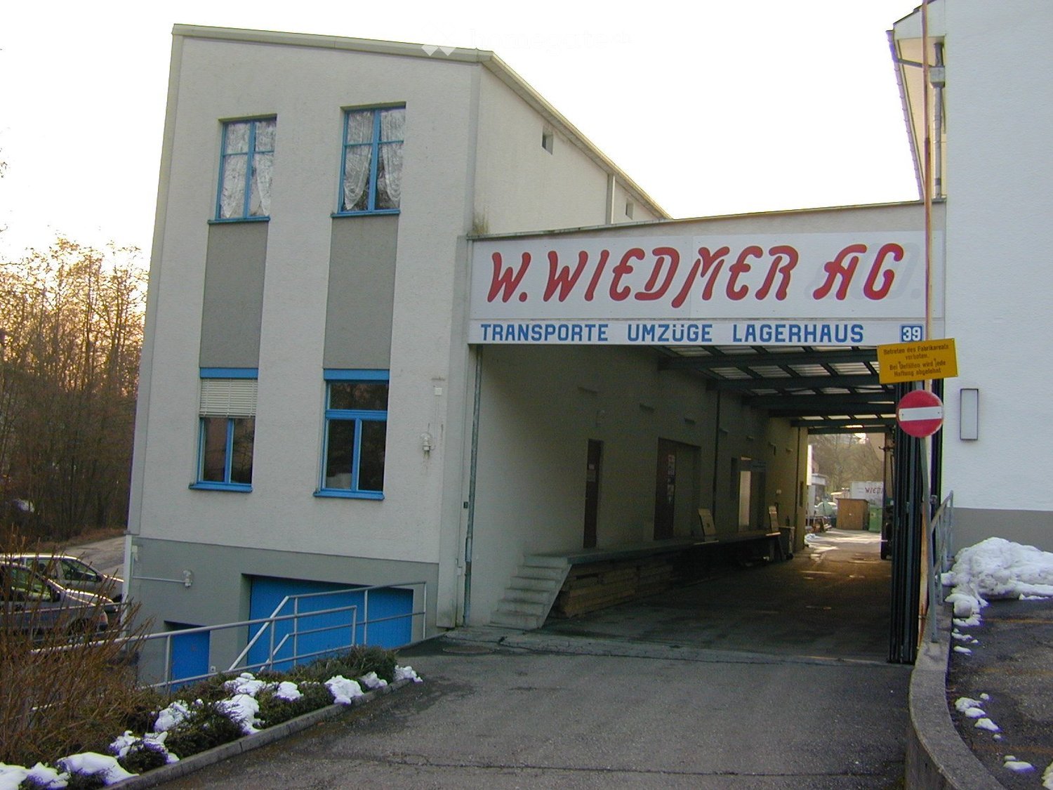 white building, blue windows, blue doors, sign reading 'W. Wiedmer AG', entrance with stairs, parking area