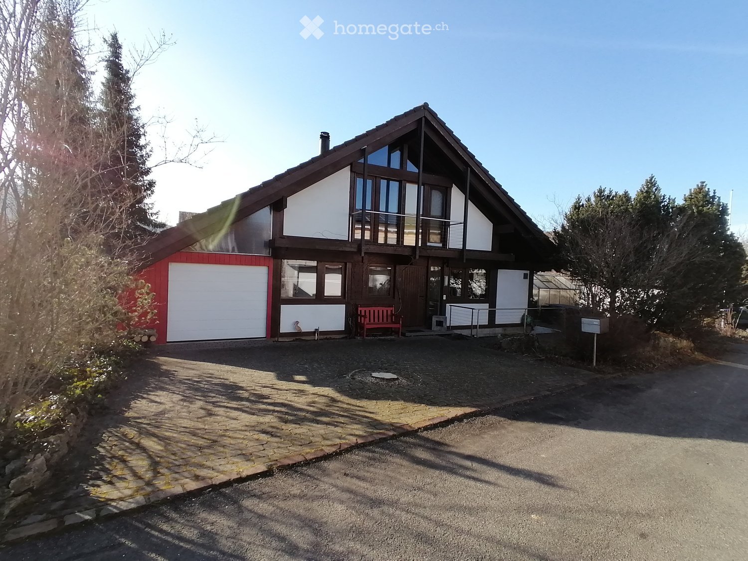Detached house, red and white exterior, two garages, red bench, paved driveway, trees, plants