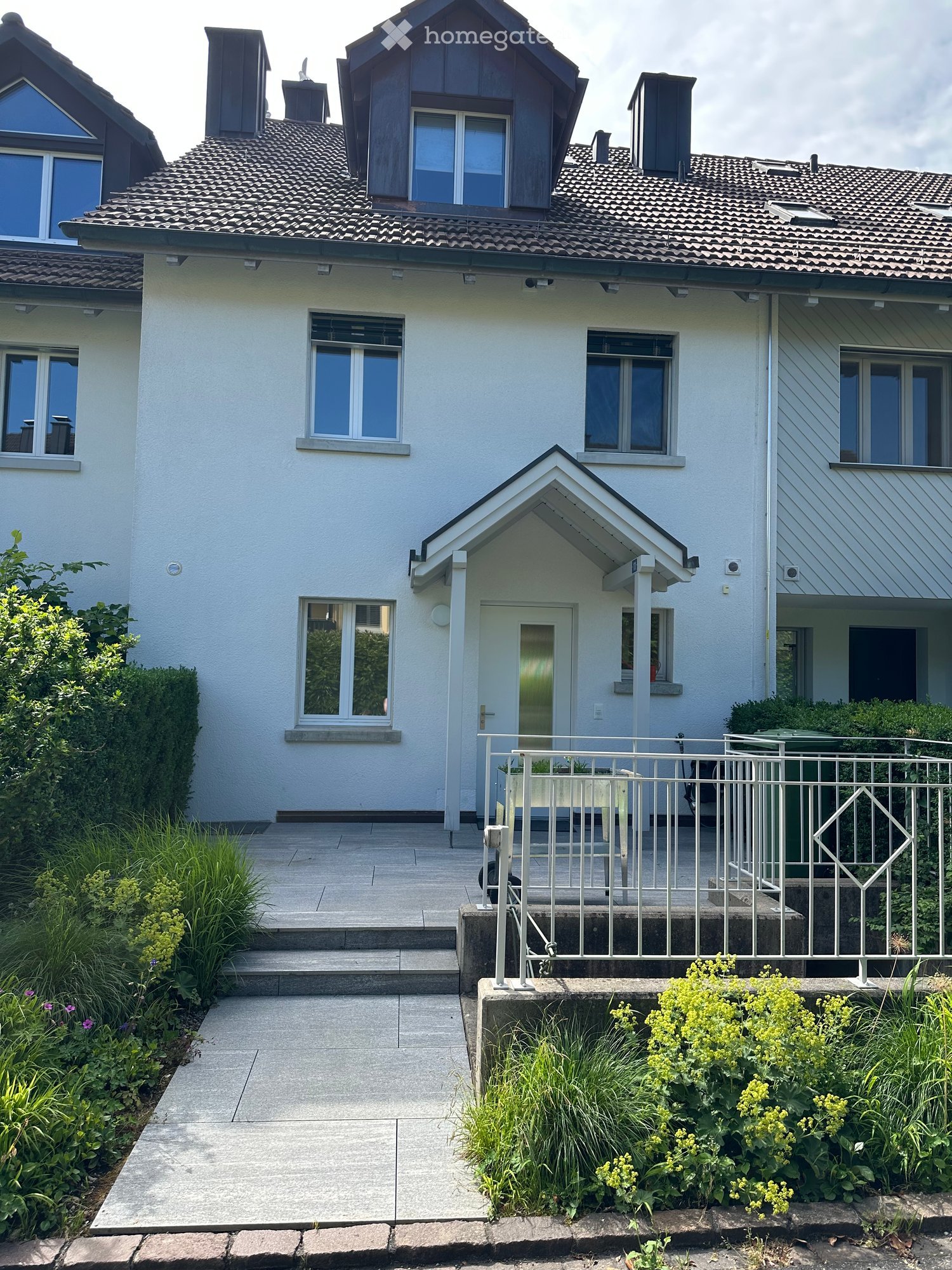 Two-story house with white exterior, multiple windows, and a small balcony area with a railing. The entrance features a small porch with steps leading up to a white door. The surrounding area includes a well-maintained garden with various plants and shrub