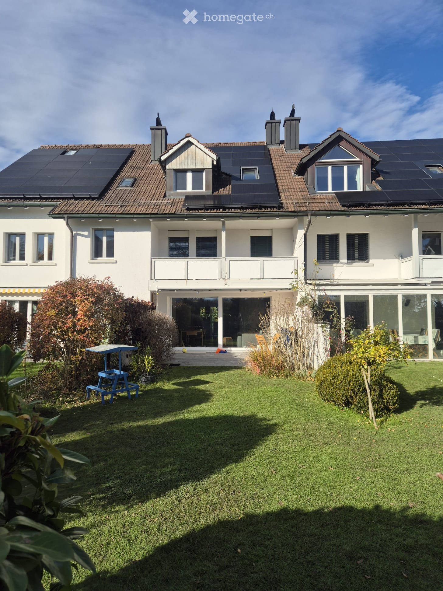 2 story house, white exterior, brown roof, solar panels, large windows, balcony, garden, blue picnic table