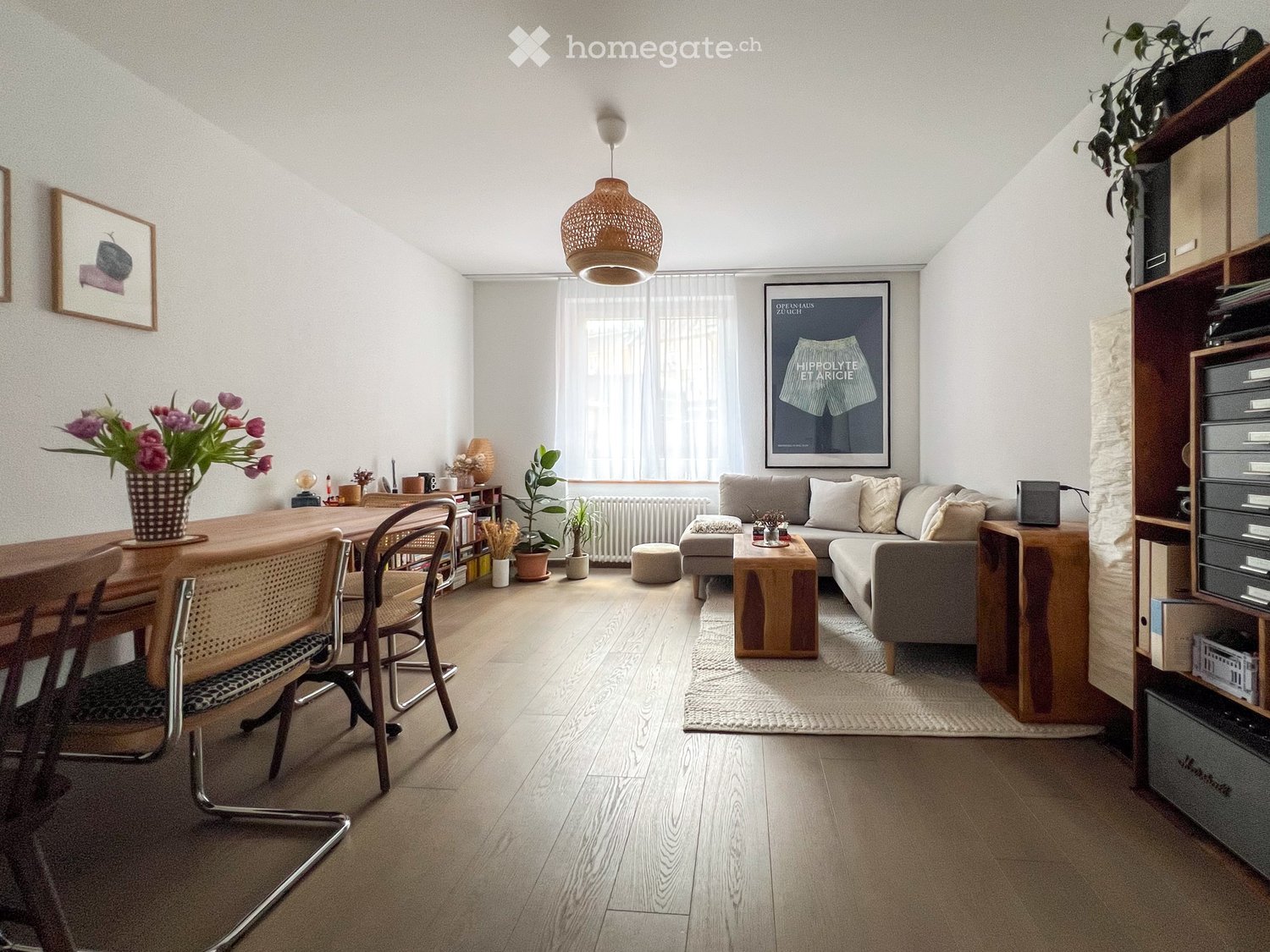 Dining area with wooden table, chairs, and flower vase. Living area with couch, coffee table, and rug. Shelves with books and plants. Picture frames on the wall.