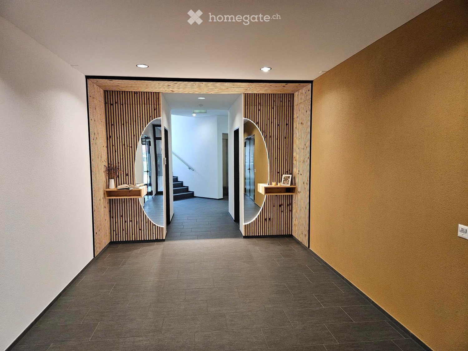 Empty corridor with tiled floor, wooden panels, two large mirrors, a staircase and decorative items on wooden shelves