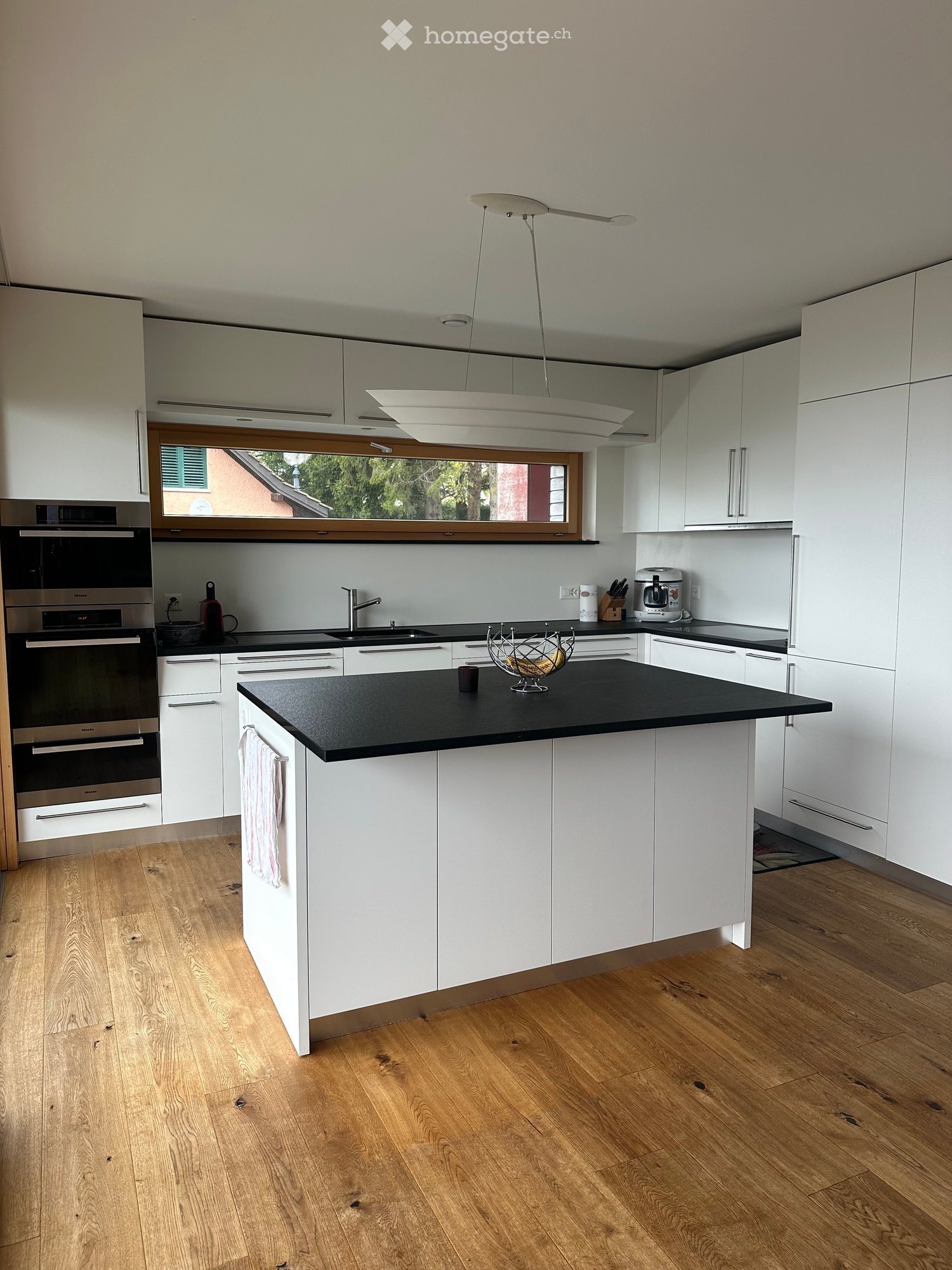 modern kitchen with white cabinets, black countertops, wooden floors, large window, hanging lamp, built-in oven, coffee machine, sink, faucet