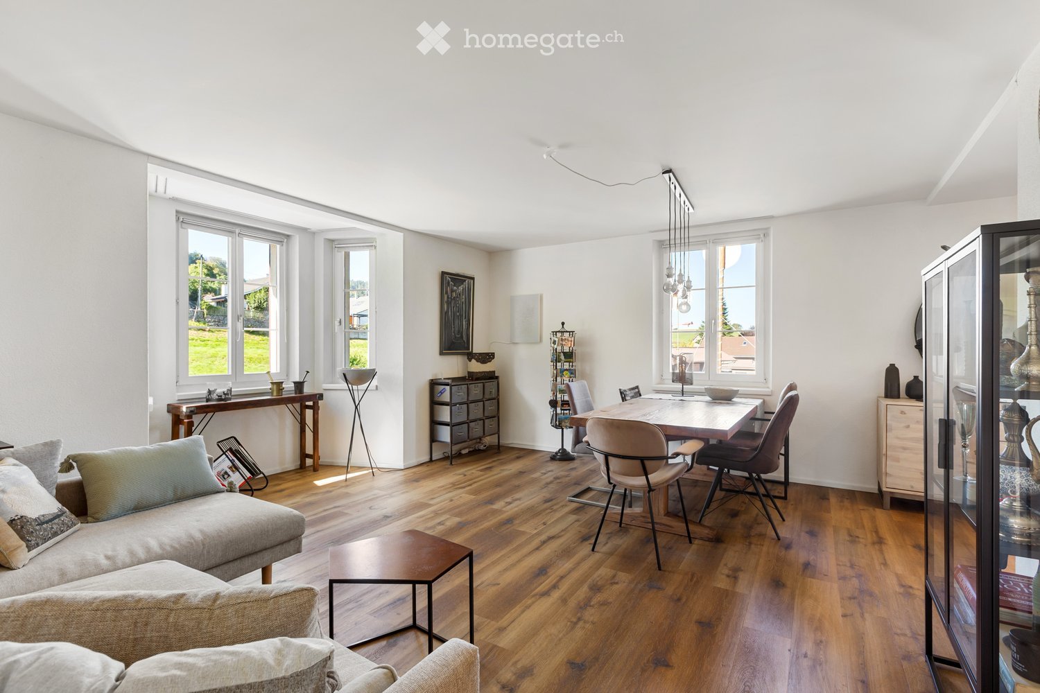 Open living and dining area with wooden floor, large windows, dining table, chairs, couch, side table, cabinet, and decorative items.