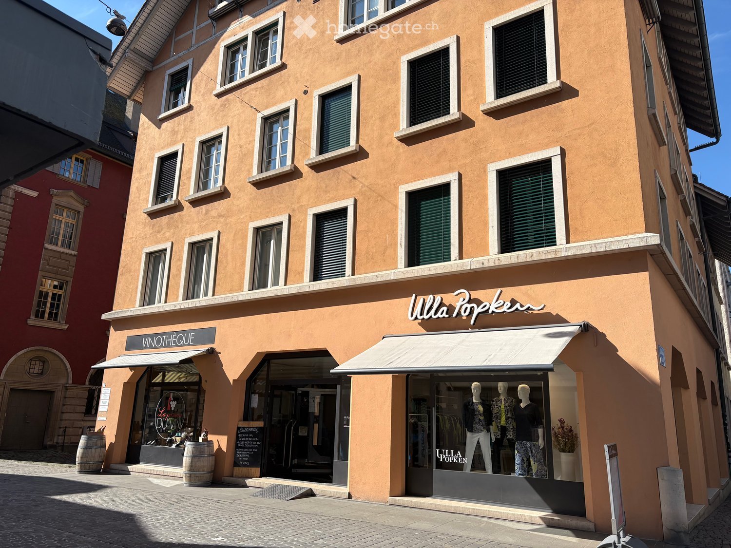 A three-story building with a beige exterior, multiple windows, and a white awning over the entrance. The ground floor has two shops with glass doors and windows, one of which has a sign reading 'VINOTHEQUE'. There are barrels and a sign outside the shop