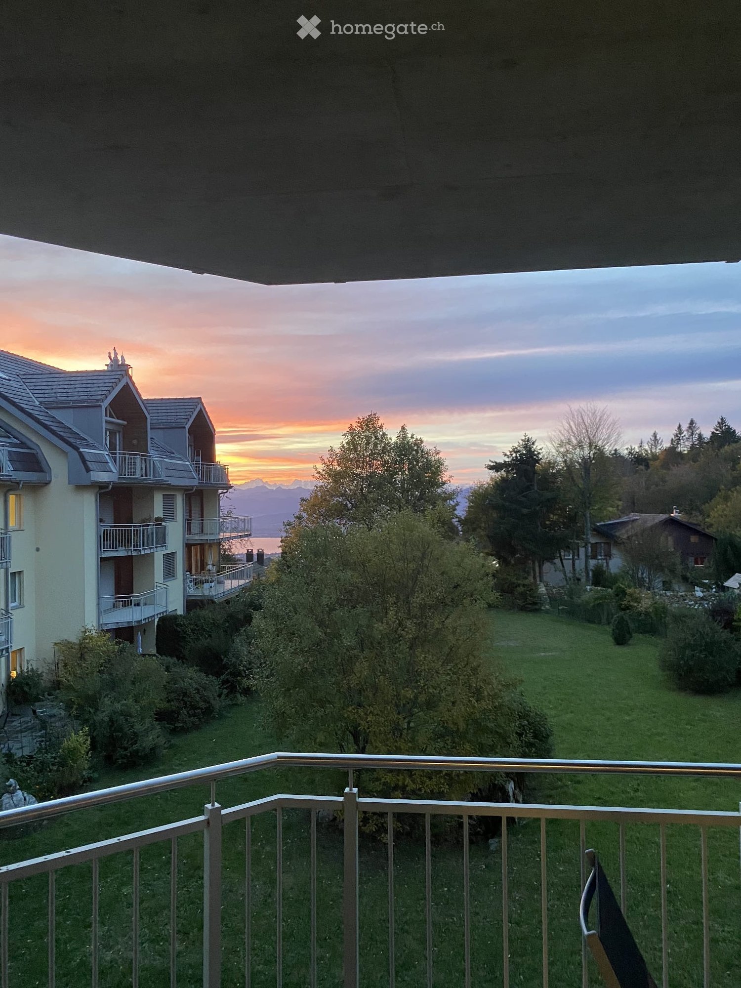 balcony with railings, overlooking a grassy area with trees and a building, sunset in the background