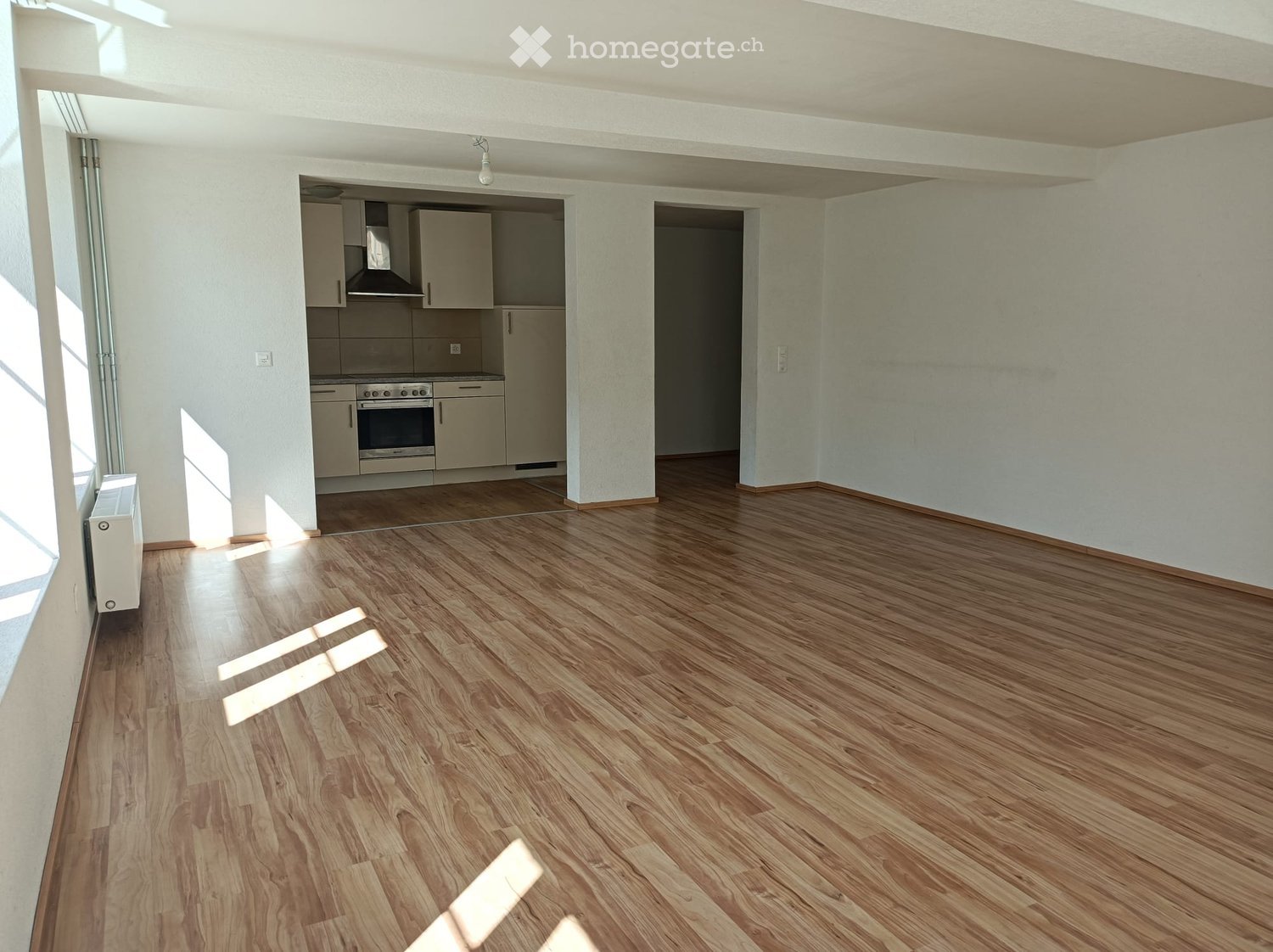 empty room, wooden floor, white walls, kitchen with stove, oven, and cabinets, windows, and a radiator