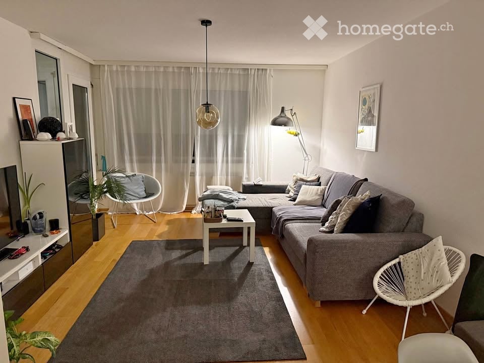 Living room with gray sofa, white coffee table, wooden floor, white walls, white curtains, pendant light, floor lamp, and various decorative items.