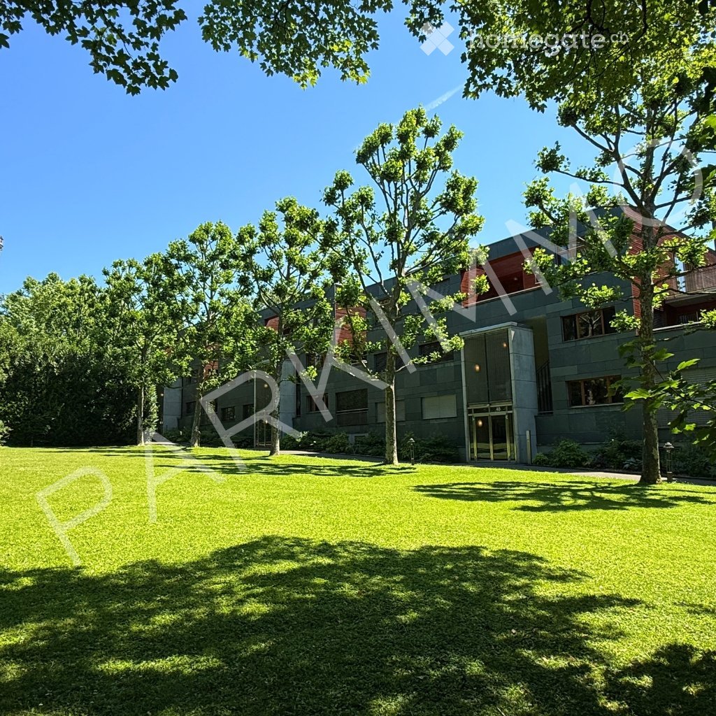 A well-maintained lawn with mature trees and a modern building in the background, featuring a clear blue sky and a peaceful setting.