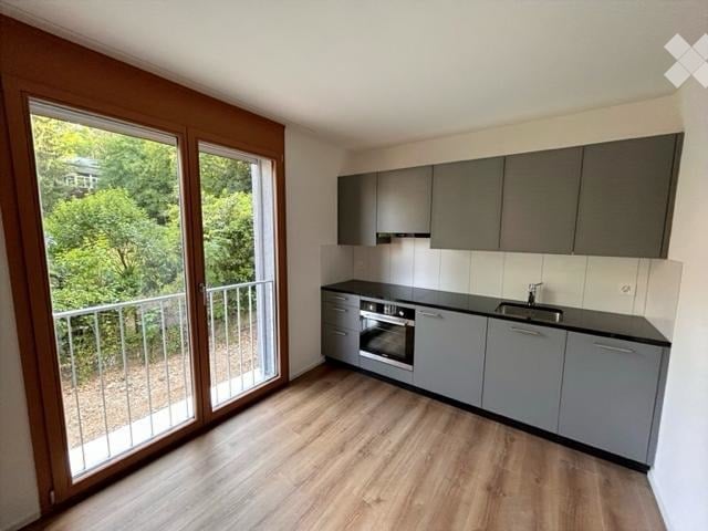 modern kitchen with wooden floor, gray cabinets, black countertops, oven, sink, sliding glass door to balcony, view of trees