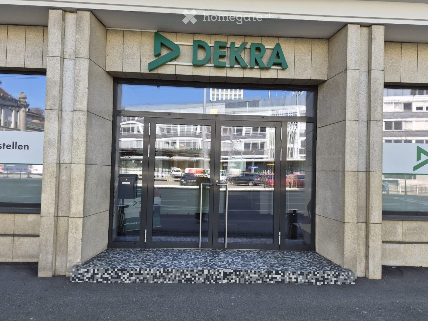 Entrance of a building with a glass door, a sign reading 'DEKRA', and a tiled floor.