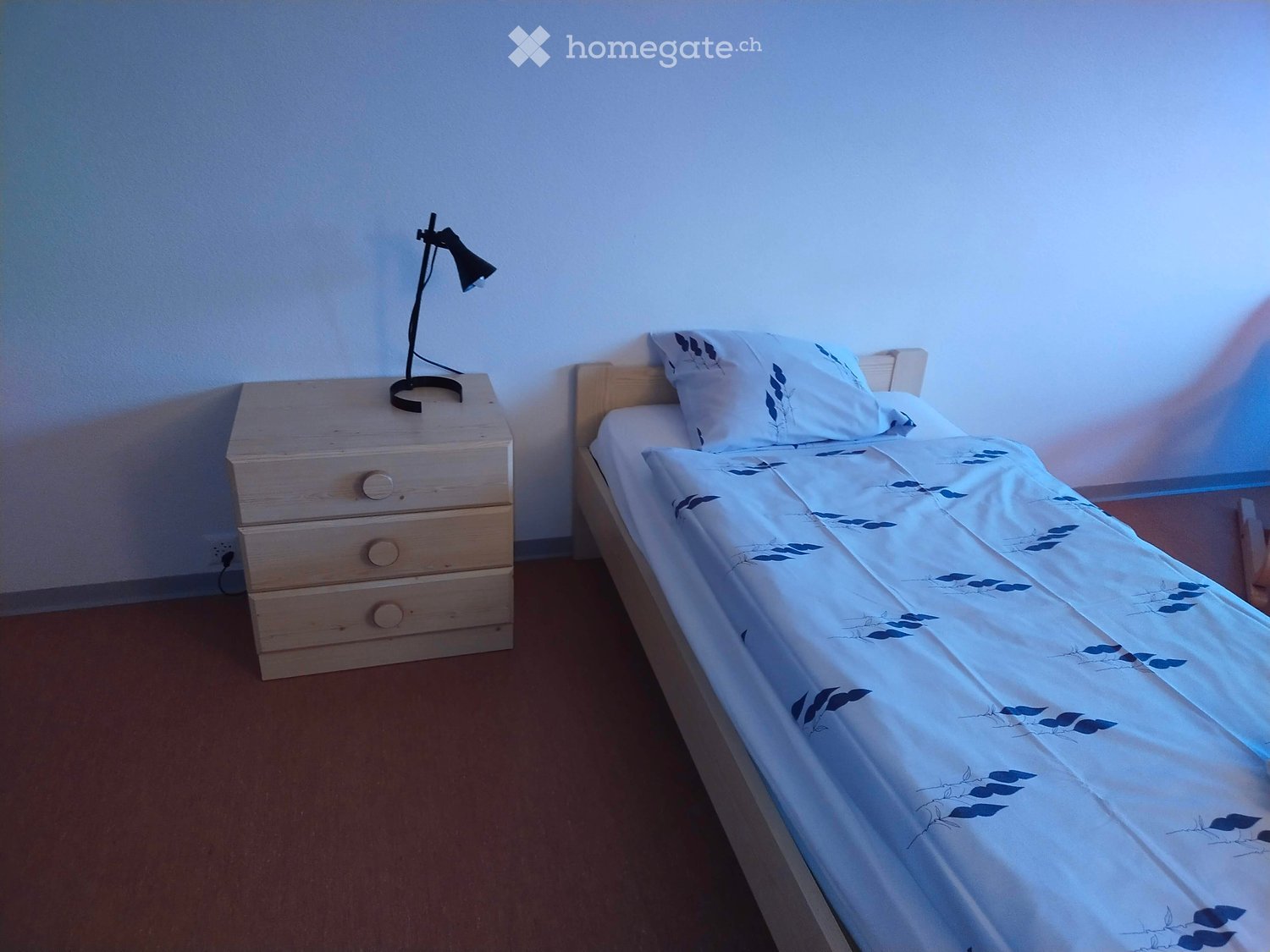 Single bed, wooden nightstand with lamp, white walls, brown floor, white bedding