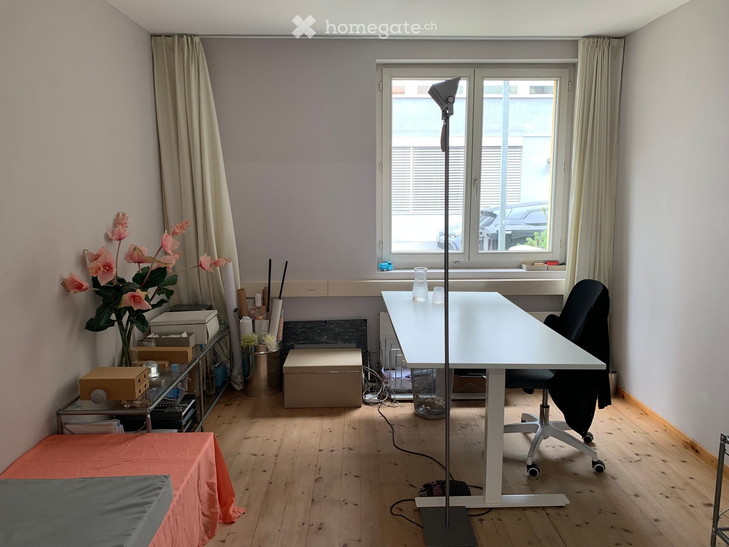 A room with a desk, chair, flowers, and a window with a view of a car.