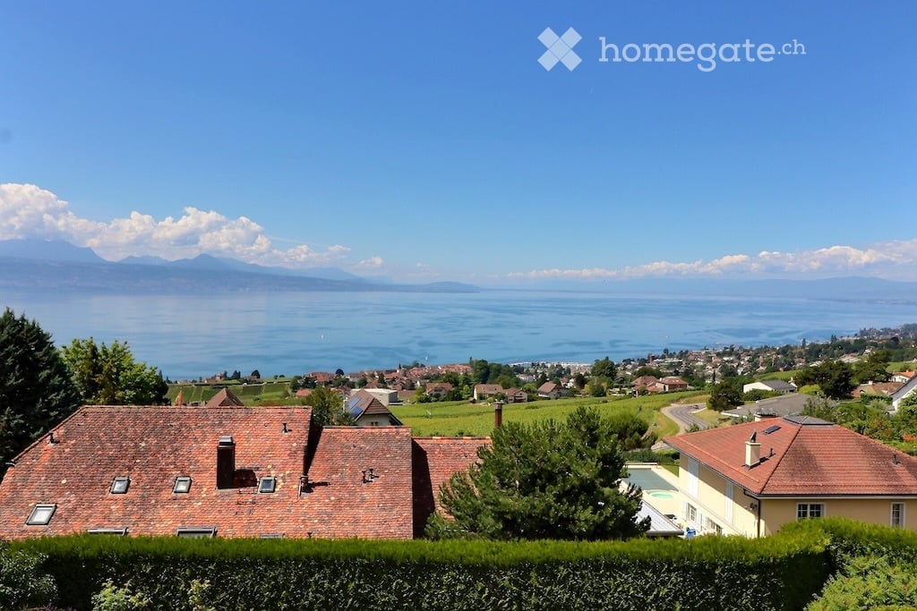 House with red roof, overlooking a lake and mountains, surrounded by greenery and a town in the distance