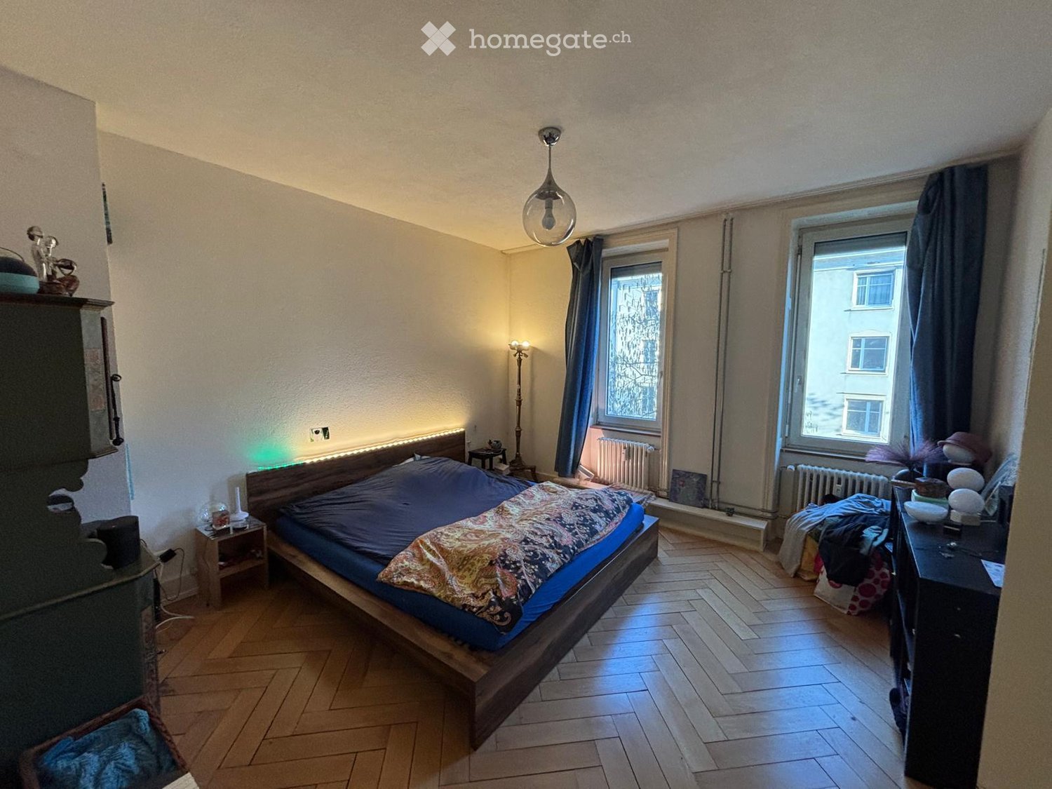 Bedroom with wooden bed, wooden floor, two windows, radiators, nightstands, lamp, and various decorative items.