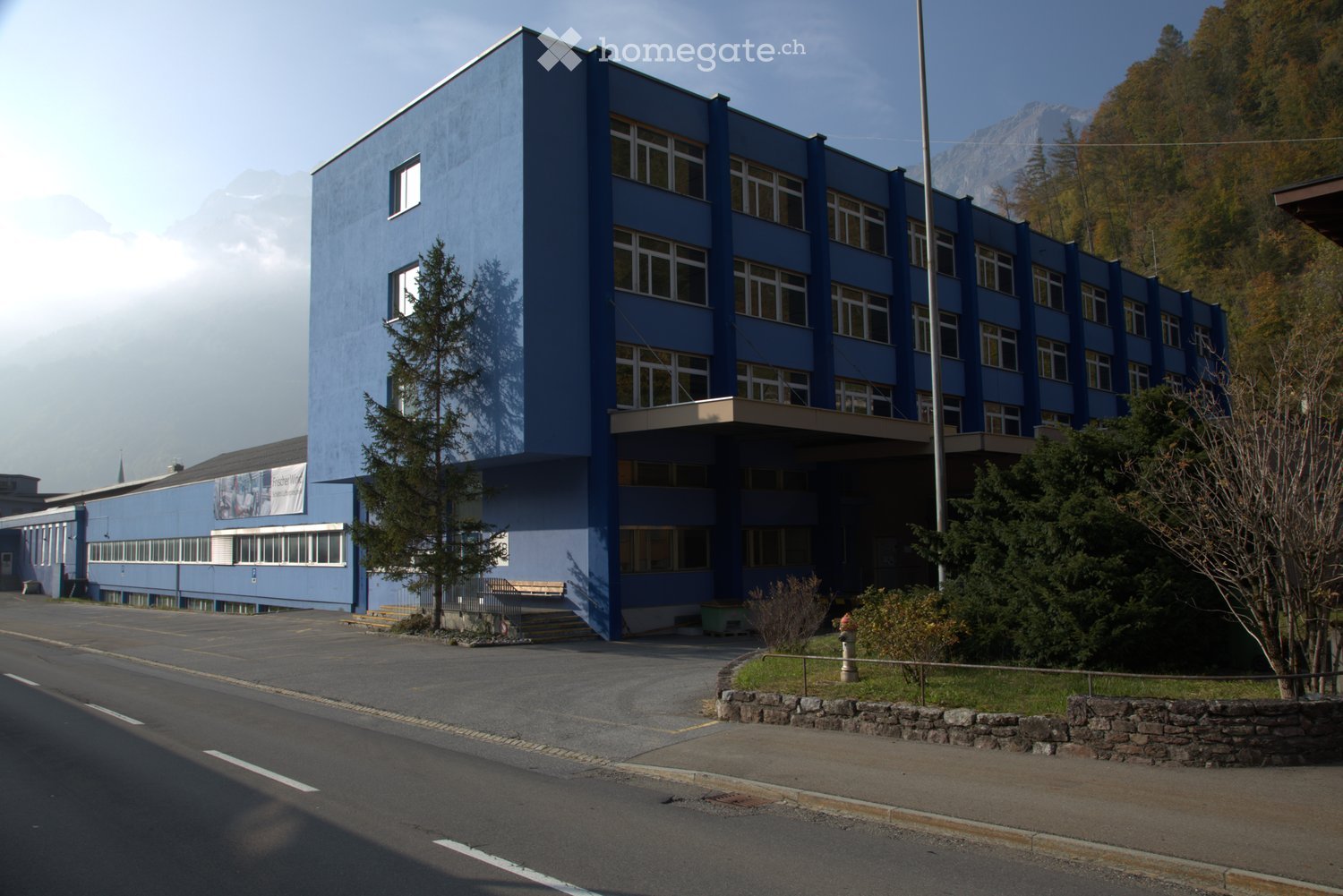 Blue building with multiple windows, located on a street with a parking lot in front, surrounded by trees and bushes, mountains in the background.