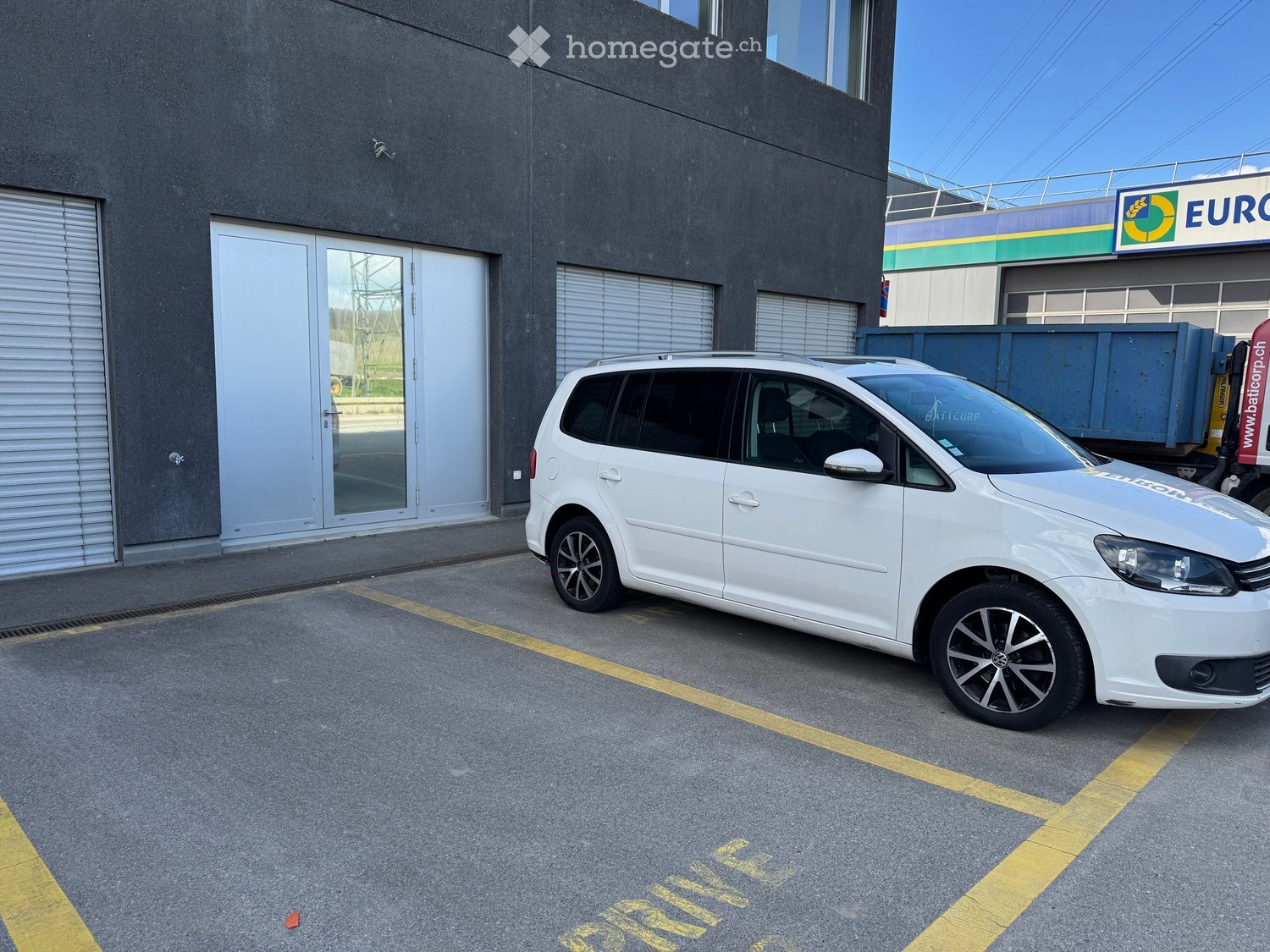 A white car parked in a parking lot in front of a building with a glass door and two garages.