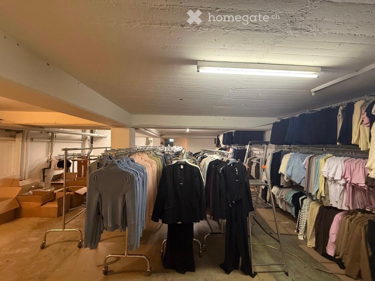 Storage room with clothes on racks, boxes on the floor, metal racks, fluorescent lights on the ceiling