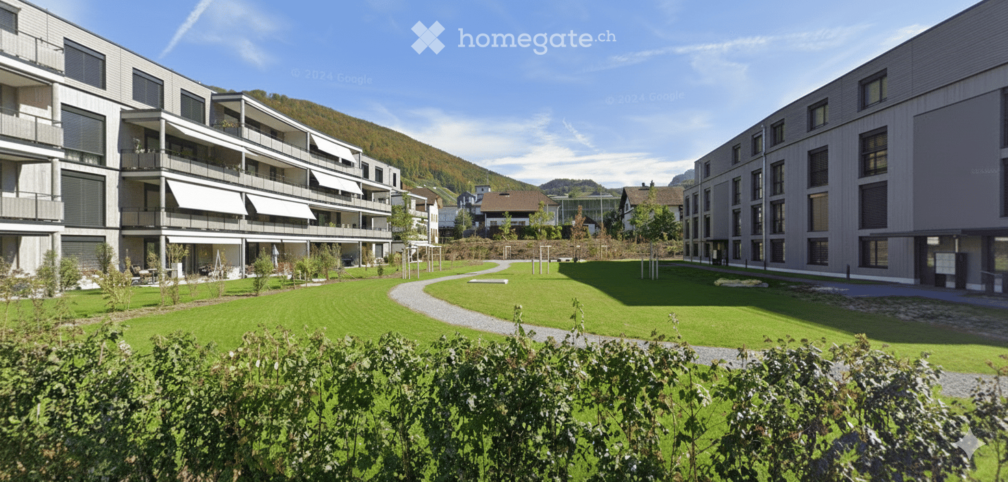 A modern, three-story apartment building with balconies, surrounded by a large garden with paths, greenery, and distant mountains. Buildings and trees are in the background.