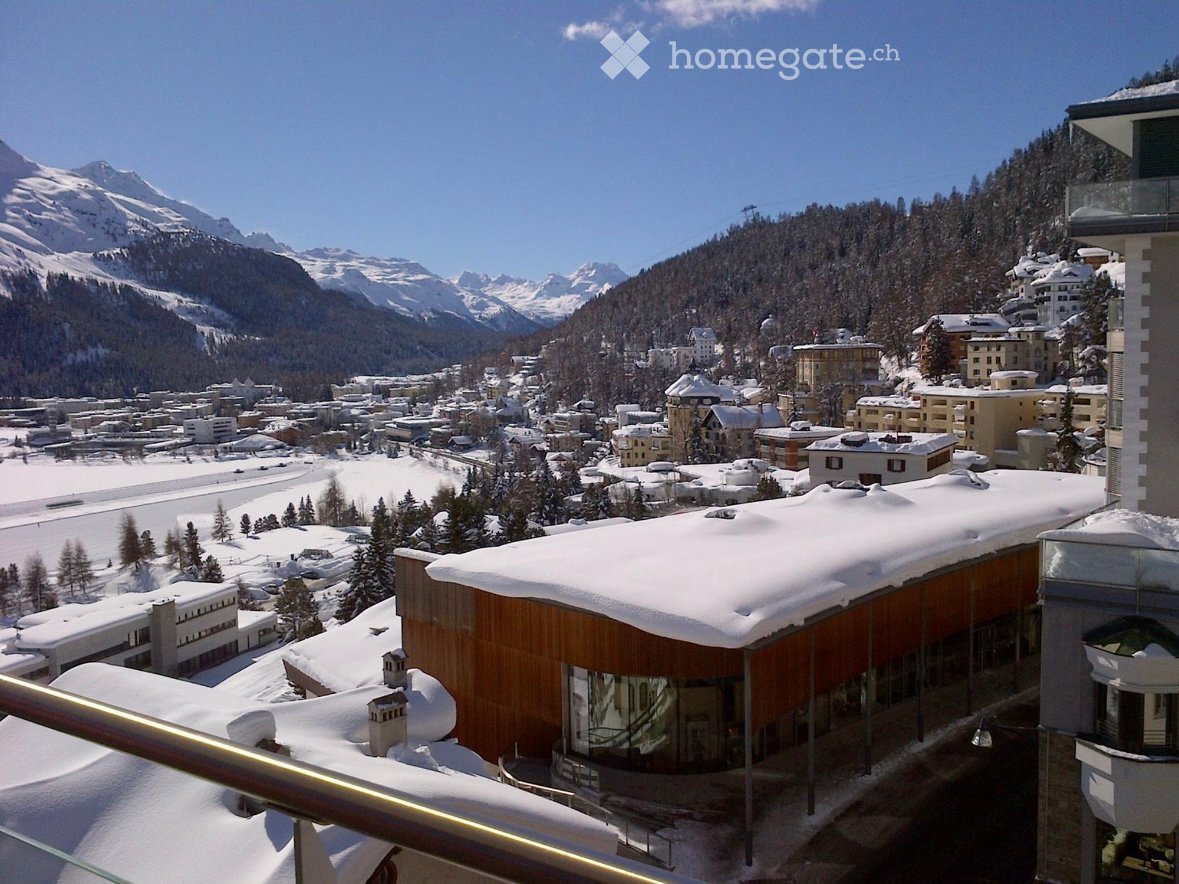 Building with a glass facade, snow-covered roof, surrounded by a snowy landscape with mountains and a town in the distance