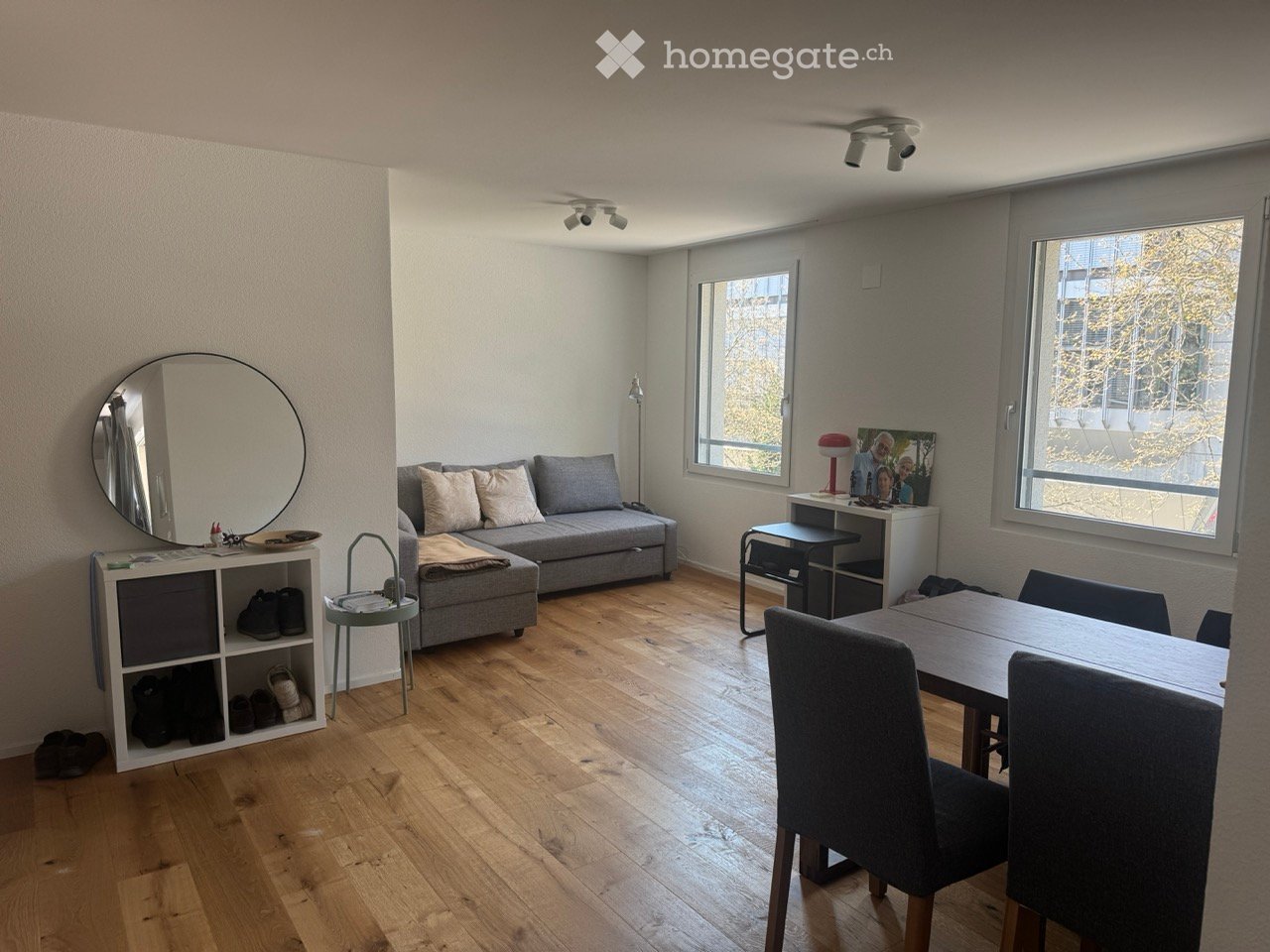 Living room with wooden floor, gray sofa, round mirror, dining table, chairs, white shelves, lamps, and windows