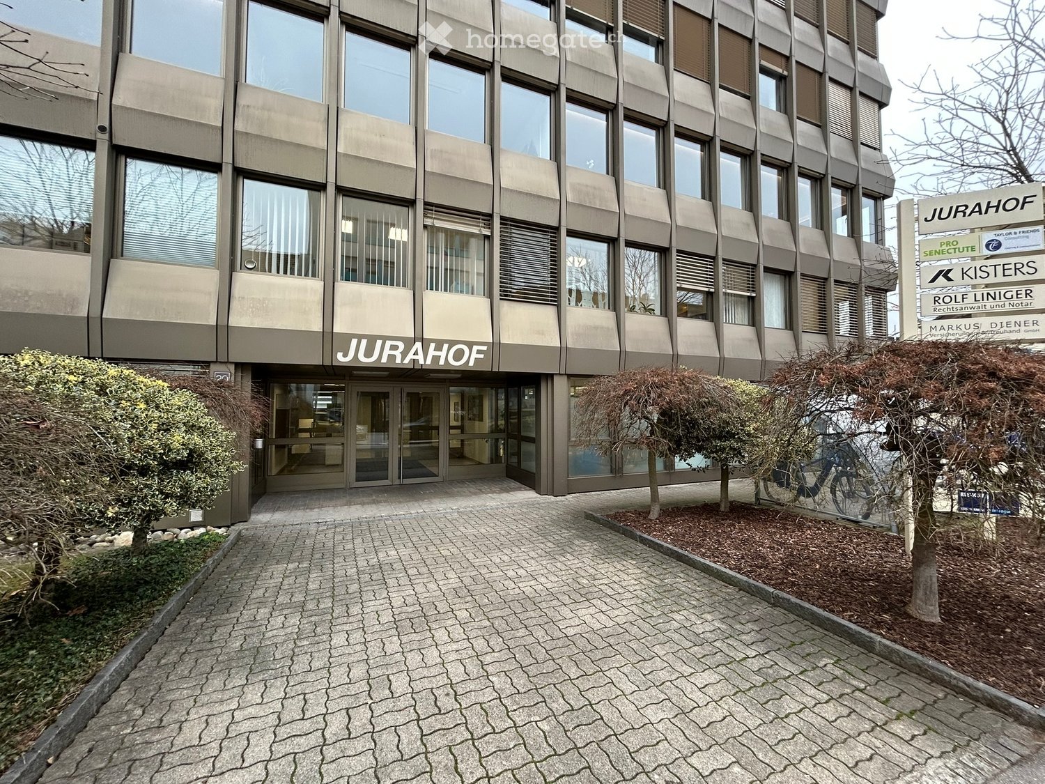 Modern office building, Jurahof, large glass windows, brick facade, entrance with glass doors, paved entrance area, greenery
