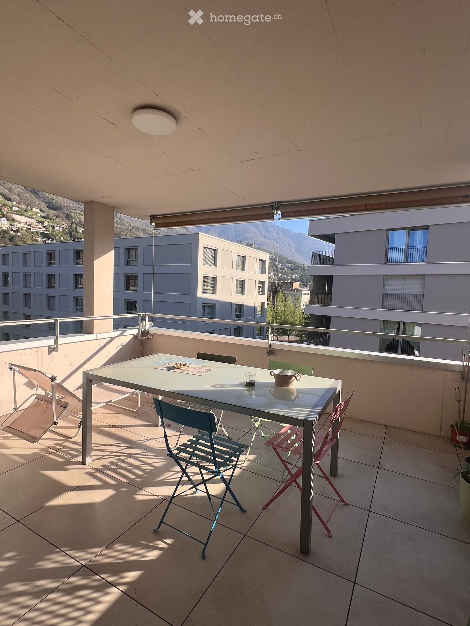 A balcony with a table and chairs, a bowl, and a glass on the table. The balcony has a metal railing and a tiled floor. There is a view of the mountains and buildings in the distance.